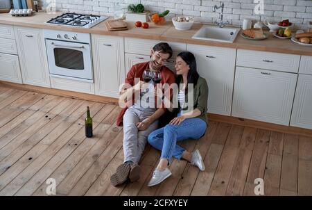 Aperitifzeit. Volle Länge der jungen schönen Paar sitzt auf dem Boden in der modernen Küche, klirrende Gläser mit Rotwein und Ausgaben groß Stockfoto