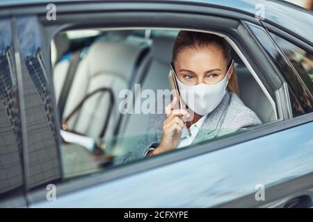 Covid 19 oder Coronavirus-Ausbruch, kaukasische Geschäftsfrau trägt medizinische Schutzmaske reden auf dem Smartphone, während auf dem Rücksitz im Auto sitzen Stockfoto