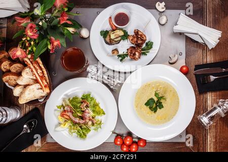 Draufsicht auf das Set Food: Spieße mit Pilzsuppe, Salat und Hähnchengrill auf einem Holztisch. Drei-Gänge-Business Lunch Stockfoto