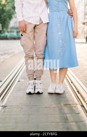 Zugeschnittenes Bild von Beinen von schönen stilvollen jungen Paar in der Liebe, Frau in blauem Kleid und Mann in beigen Hosen, auf dem Bürgersteig Straße und Straßenbahn-Spur stehen Stockfoto