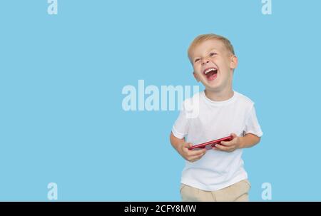 Niedliche smilling Kind Junge mit Smartphone auf blauem Hintergrund. Werbung für pädagogische Anwendungen auf einem Handy für Kinder. Lustiges Gesicht. Stockfoto