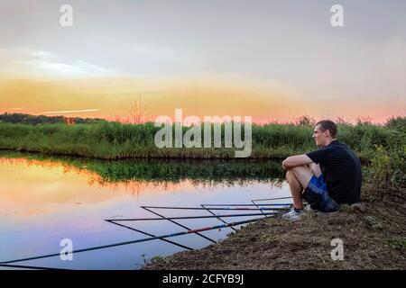 Ruhiger entspannter Mann fängt Angeln auf Angelruten sitzen auf Das Ufer des Sees bei Sonnenuntergang Stockfoto