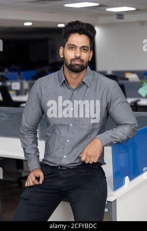 Porträt eines gutaussehenden jungen indischen Geschäftsmannes, der lässig gegen seinen Arbeitsplatz steht. Unternehmensumgebung. Stockfoto