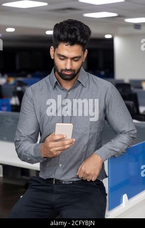 Portrait eines hübschen jungen indischen Geschäftsmannes, der sein Telefon auf E-Mail und soziale Medien überprüft. Unternehmensumgebung. Stockfoto