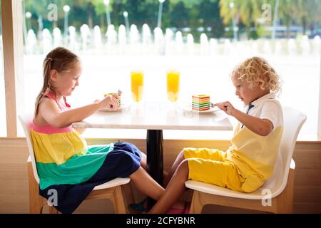 Kinder essen Regenbogenkuchen im Restaurant. Junge und Mädchen mit Süßigkeiten und Gebäck. Kinder trinken frischen Orangensaft im Café. Familie, die im Stadtcafé auswärts isst Stockfoto