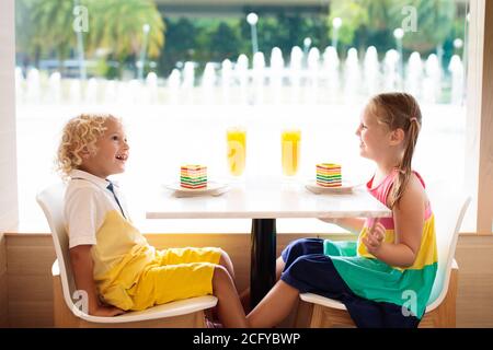 Kinder essen Regenbogenkuchen im Restaurant. Junge und Mädchen mit Süßigkeiten und Gebäck. Kinder trinken frischen Orangensaft im Café. Familie, die im Stadtcafé auswärts isst Stockfoto