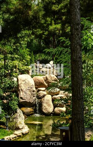 Große Steinblöcke auf einem kleinen Fluss schufen eine Wasserfall und ein kleiner Teich unter Bäumen in der "Vogel World' Park in Teheran Stockfoto