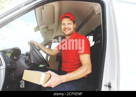 Lächelnder Fahrer des Transportservice in roter Uniform sitzt im Van Mit Box in der Hand