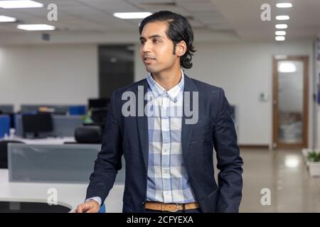 Portrait eines hübschen jungen indischen Geschäftsmannes, der von der Kamera wegschaut und vor dem Hintergrund eines Firmenbüros steht. Stockfoto