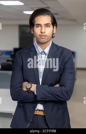 Porträt eines gutaussehenden jungen indischen Geschäftsmannes, der mit gekreuzten Armen vor dem Hintergrund eines Unternehmensbüros steht. Stockfoto