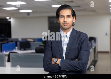 Porträt eines gutaussehenden jungen indischen Geschäftsmannes, der mit gekreuzten Armen vor dem Hintergrund eines Unternehmensbüros steht. Stockfoto