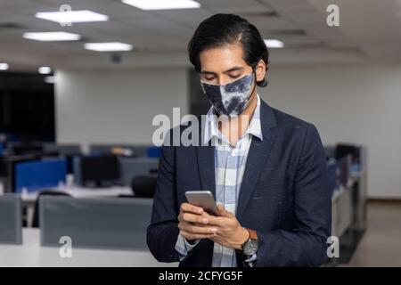 Portrait eines hübschen jungen indischen Geschäftsmannes, der Covid Schutzmaske trägt und sein Handy überprüft. Unternehmensumgebung. Stockfoto
