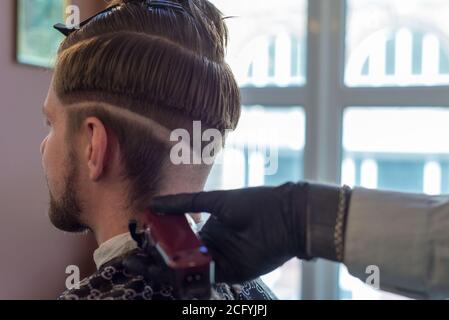 Ein Friseur schneidet einen bärtigen jungen Kerl mit einem Haarschneider und kämmt die Haare auf seinem Kopf. Die Arbeit des Meisters im Männerhaarschnitt in einem Friseurladen. Die Ba Stockfoto