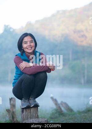 Eine asiatische Dame hockt auf einem Holzpfahl in der Nähe des Sees im Park. Stockfoto