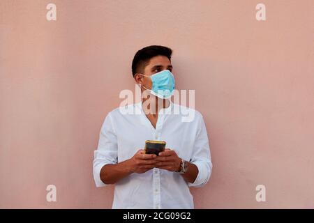Junger latein mit Maske schaut auf sein Telefon Rosa Hintergrund Stockfoto
