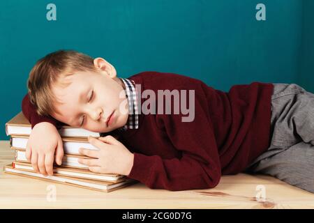 Müde Schuljunge schläft auf Büchern. Kleine Schüler schlafen auf Lehrbüchern. Kind in Schuluniform liegt auf dem Tisch mit großen Stapel von Büchern gegen Blau Stockfoto