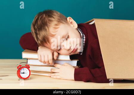 Müde Schuljunge schläft auf Büchern. Kleine Schüler schlafen auf Lehrbüchern. Kind in Schuluniform liegt auf dem Tisch mit großen Stapel von Büchern gegen Blau Stockfoto