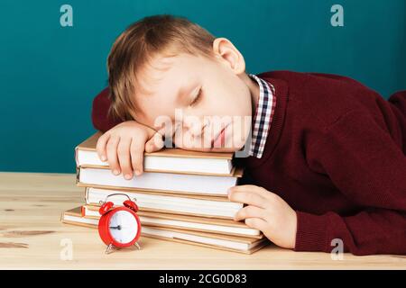 Müde Schuljunge schläft auf Büchern. Kleine Schüler schlafen auf Lehrbüchern. Kind in Schuluniform liegt auf dem Tisch mit großen Stapel von Büchern gegen Blau Stockfoto