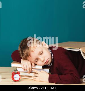 Schlafender Schuljunge. Kleiner Schüler, der auf Lehrbüchern schläft. Kind in Schuluniform liegt auf dem Tisch mit großem Bücherstapel an der blauen Wand. Augen Klo Stockfoto