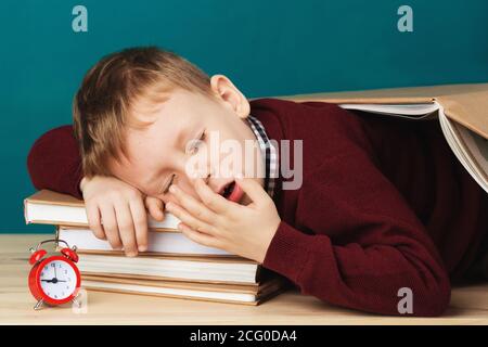 Müde Schuljunge schläft auf Büchern. Kleine Schüler schlafen auf Lehrbüchern. Kind in Schuluniform liegt auf dem Tisch mit großen Stapel von Büchern gegen Blau Stockfoto