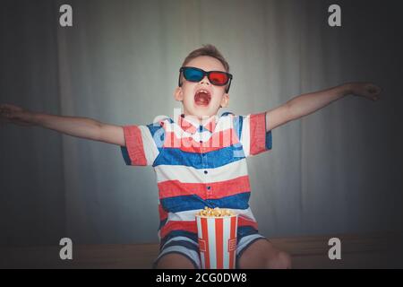 Kleiner Junge schaut einen Film in 3D-Brille im Kino oder zu Hause. Kleines Kind essen Popcorn auf grauem Hintergrund. Home Entertainment-System. Niedliches Kind im Vintage-Zimt Stockfoto