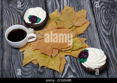 Schokoladen-Muffins mit Sahne Blume. Eine Tasse schwarzen Kaffee. Unter den getrockneten Ahornblättern. Liegt auf Kiefernbrettern schwarz und weiß lackiert. Stockfoto