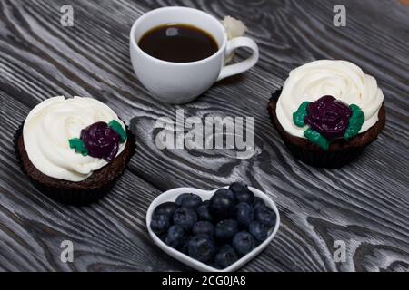Schokoladen-Muffins mit Sahne Blume. Eine Tasse schwarzen Kaffee. Untertasse mit Heidelbeeren. Liegt auf Kiefernbrettern schwarz und weiß lackiert. Stockfoto