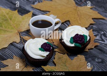 Schokoladen-Muffins mit Sahne Blume. Eine Tasse schwarzen Kaffee. Unter den getrockneten Ahornblättern. Liegt auf Kiefernbrettern schwarz und weiß lackiert. Stockfoto