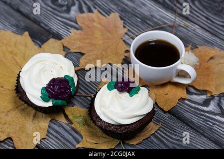 Schokoladen-Muffins mit Sahne Blume. Eine Tasse schwarzen Kaffee. Unter den getrockneten Ahornblättern. Liegt auf Kiefernbrettern schwarz und weiß lackiert. Stockfoto
