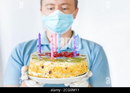 Ein Kerl in einer Maske und Handschuhen hält einen Kuchen in der Hand. Geburtstagsfeier während covid-19 Quarantäne. Stockfoto