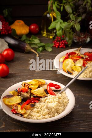 Porridge Couscous mit gebackenem Gemüse, Zucchini und Zwiebeln, Paprika und Auberginen mit Gewürzen, Teller mit Lebensmitteln auf dunklem Holzgrund Stockfoto