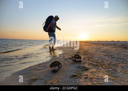 Sandalen auf nassem Sand am Meer. Der Typ läuft mit einem Rucksack entlang der Küste. Die Strahlen der Abendsonne spiegeln sich im nassen Sand Stockfoto