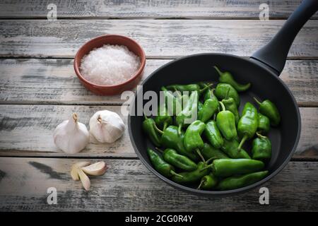 Eine schwarze Pfanne gefüllt mit rohen Pimentos, schön angeordnet auf einem weißen schäbig chic Holzteller, daneben eine Schüssel mit grobem Meersalz und Knoblauch Stockfoto