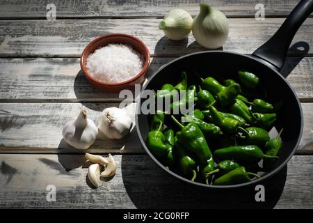 Eine schwarze Pfanne gefüllt mit rohen Pimentos, schön angeordnet auf einem weißen schäbig chic Holzteller, daneben eine Schüssel mit grobem Meersalz und Knoblauch Stockfoto