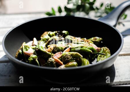 Eine schwarze Pfanne gefüllt mit frittierten Pimentos, schön arrangiert auf einem weißen schäbig schicken Holzteller, daneben eine Schüssel mit grobem Meersalz und Knoblauch Stockfoto