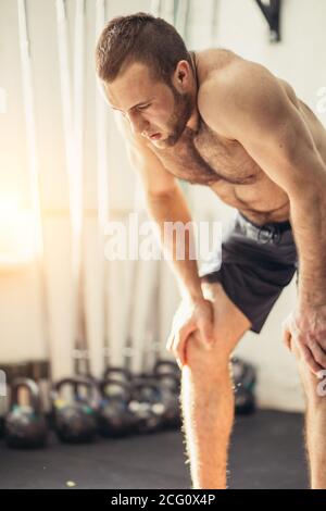 Aufnahme eines jungen Mannes in Sportkleidung, der sich mit den Händen auf den Knien im Fitnessstudio beugte. Fitness-Mann, der nach intensivem Cross-Fit-Training müde aussieht Stockfoto