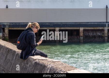 Eine junge Frau, die an einer Wand am Meer sitzt Ihr Mobiltelefon oder Mobiltelefon betrachten Stockfoto