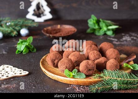 Dunkle Schokoladentrüffel mit rohem Kakaopulver, Minze und Tannenzweigen, dunkelbrauner Hintergrund. Weihnachtskomposition. Stockfoto