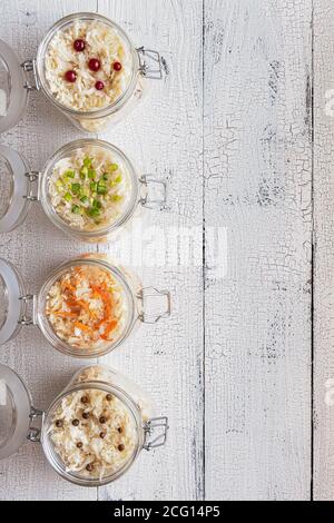 Vier Gläser voll mit hausgemachtem fermentiertem Kohl mit Karotte, Frühlingszwiebeln, schwarzem Pfeffer und Preiselbeeren auf vertikalem weißen Holzhintergrund. Flach liegend Stockfoto