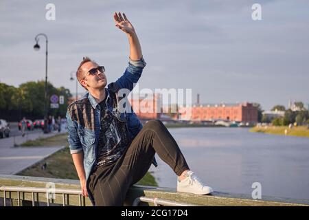 Der junge Mann winkt jemandem auf dem Hintergrund des historischen Zentrums von Sankt Petersburg, Russland, die Hand zu. Stockfoto