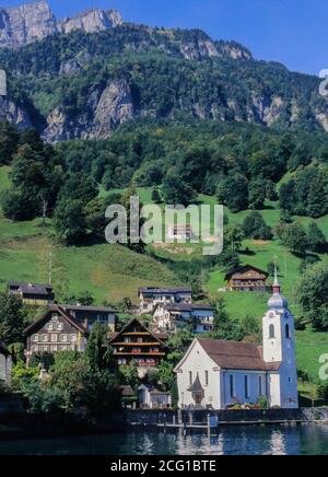 Weiße Dorfkirche St. Idda am Ufer des Vierwaldstättersees mit Bergweiden und Felsvorsprüngen im Hintergrund hinter dem Seedorf Bauen, Stockfoto