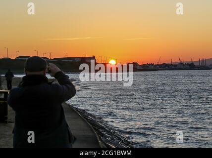 Mann, der am Handy den Sonnenuntergang über der Stadt Belfast mit der Silhouette der Belfast Werft hinter dem Belfast Lough in Holywood fotografiert. Stockfoto