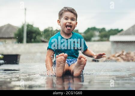 Vorderansicht Foto eines kaukasischen Jungen gestikuliert Glück mit Zunge beim Spielen barfuß im Wasser auf dem Boden nach Der Regen Stockfoto