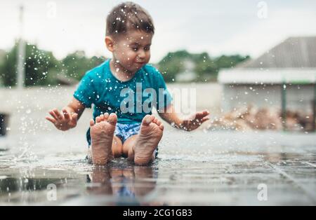 Kaukasischer kleiner Junge in blauer Kleidung spielt barfuß hinein Das Wasser auf dem Boden macht Tropfen mit Handflächen Stockfoto