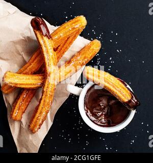 Churros mit Zucker in Schokoladensauce auf schwarzem Hintergrund getaucht. Churro-Sticks. Gebratenes Teiggebäck, Draufsicht Stockfoto