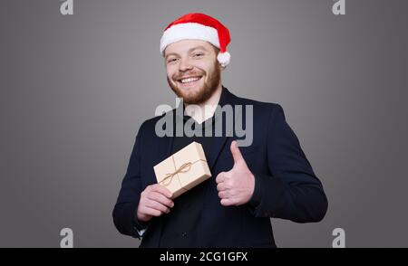 Bärtiger Unternehmer trägt einen weihnachtsmütze posiert in einem Schwarzer Anzug mit einem Geschenk gestikuliert das Like Zeichen an Eine graue Studiowand Stockfoto