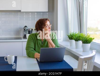 Durchdachte ältere Frau, die von zu Hause aus mit einem Laptop arbeitet. Sie schaut weg. Stockfoto