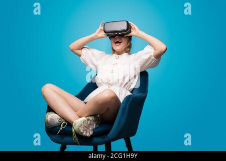 Blonde Dame im Sessel sitzt mit Virtual-Reality-Brille Und Spaß an einer blauen Studiowand Stockfoto