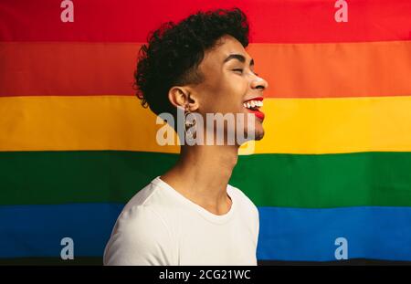 Lächelnd junge Homosexuell Mann mit Make-up steht gegen Stolz Flagge. Mann mit rotem Lippenstift und Ohrring lachend vor der Regenbogenfahne von Homosexuell Stolz. Stockfoto