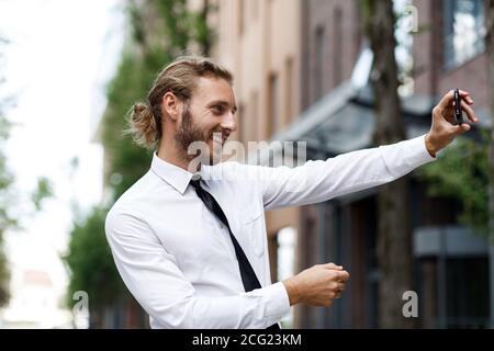 Porträt eines erfolgreichen Geschäftsmannes, der ein Selfie und ein Lächeln nimmt. Ein lockiger Manager, in einem weißen Hemd und Krawatte, nimmt glücklich ein Selfie auf einem Telefon Stockfoto